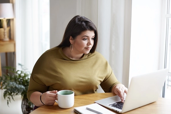 A young woman wearing elegant sweater and round earrings works in front of open laptop.